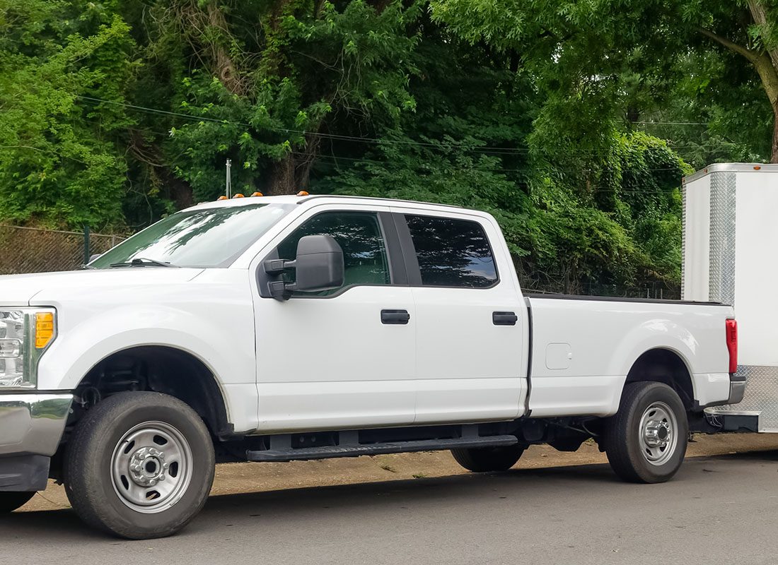 Auto Insurance - View of a Parked White Truck with an Attached Trailer on a Country Road Surrounded by Green Foliage