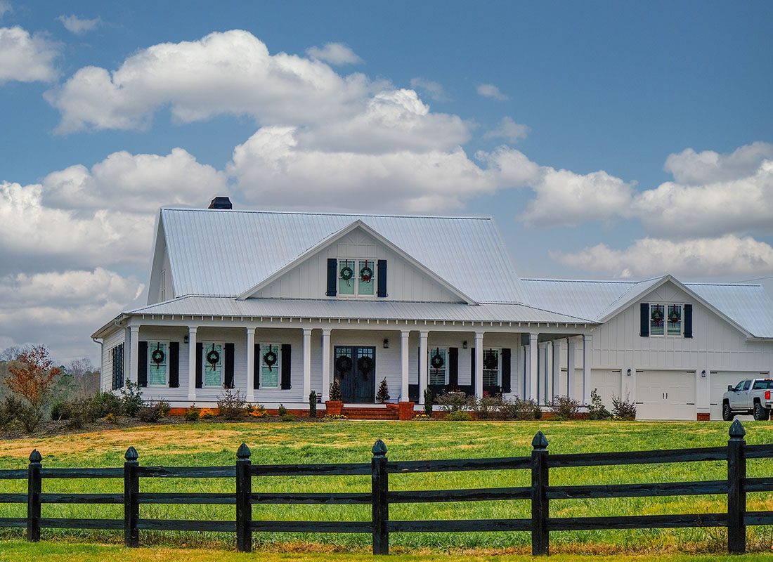 Home Insurance - Exterior View of a Two Story Barn Style Home with a Large Front Porch and Three Car Garage Against a Cloudy Blue Sky with a Wooden Fence Around the Front Yard
